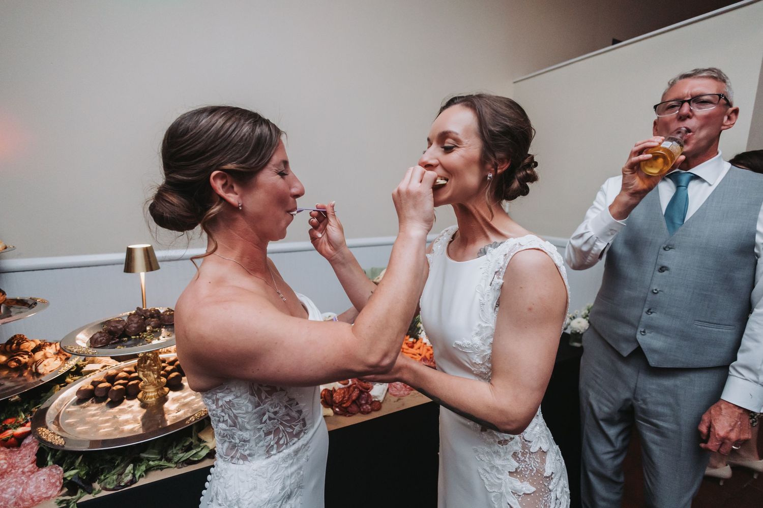 One bride feeds the other a bite of food while a man in a suit drinks nearby at a wedding reception.