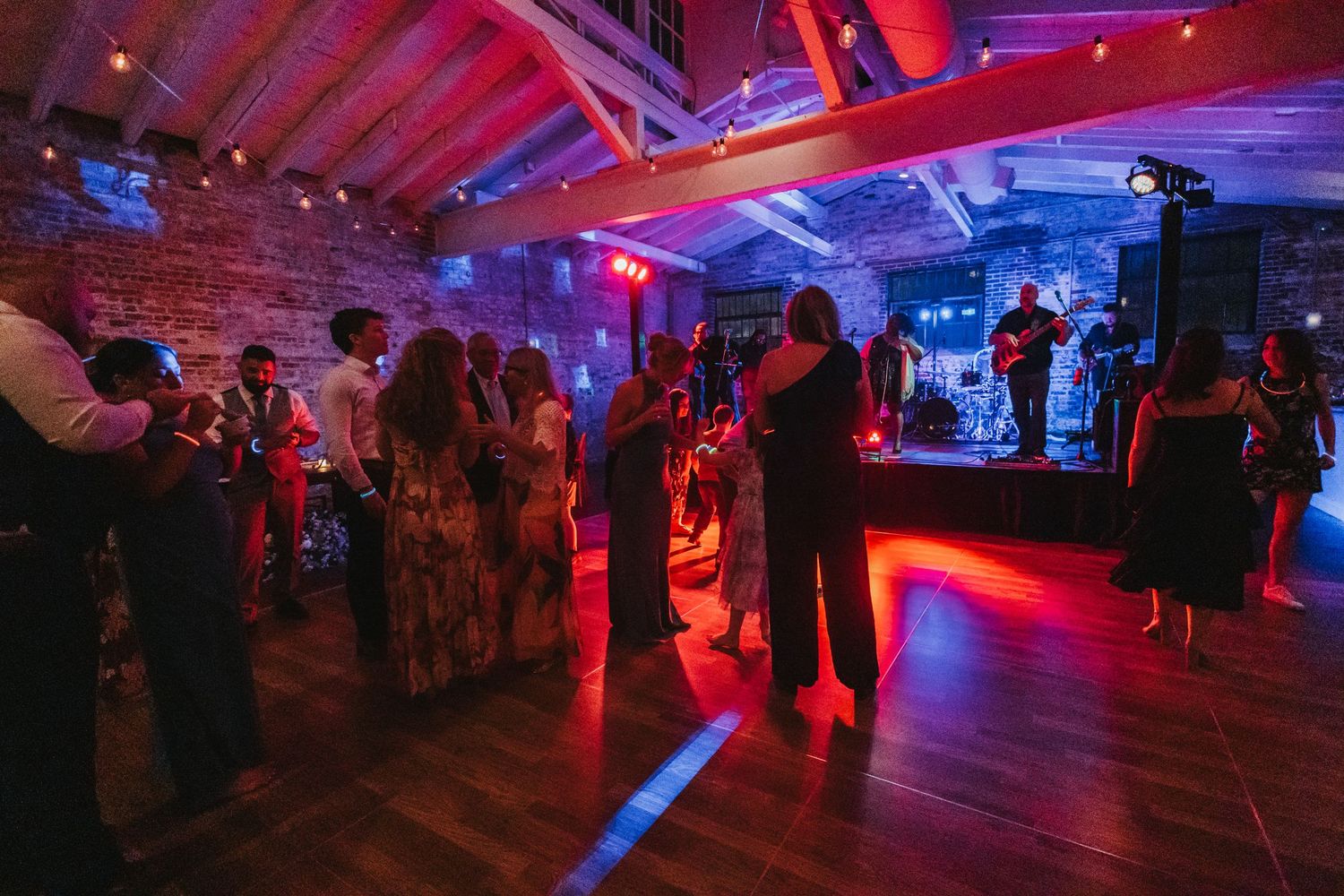 Guests dancing at a wedding reception in a rustic brick venue with red and blue stage lighting and a live band performing.