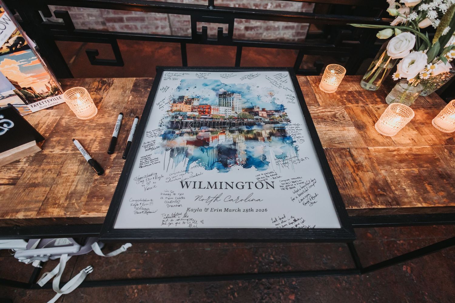 A framed watercolor print of Wilmington North Carolina sits on a rustic table with guest signatures.