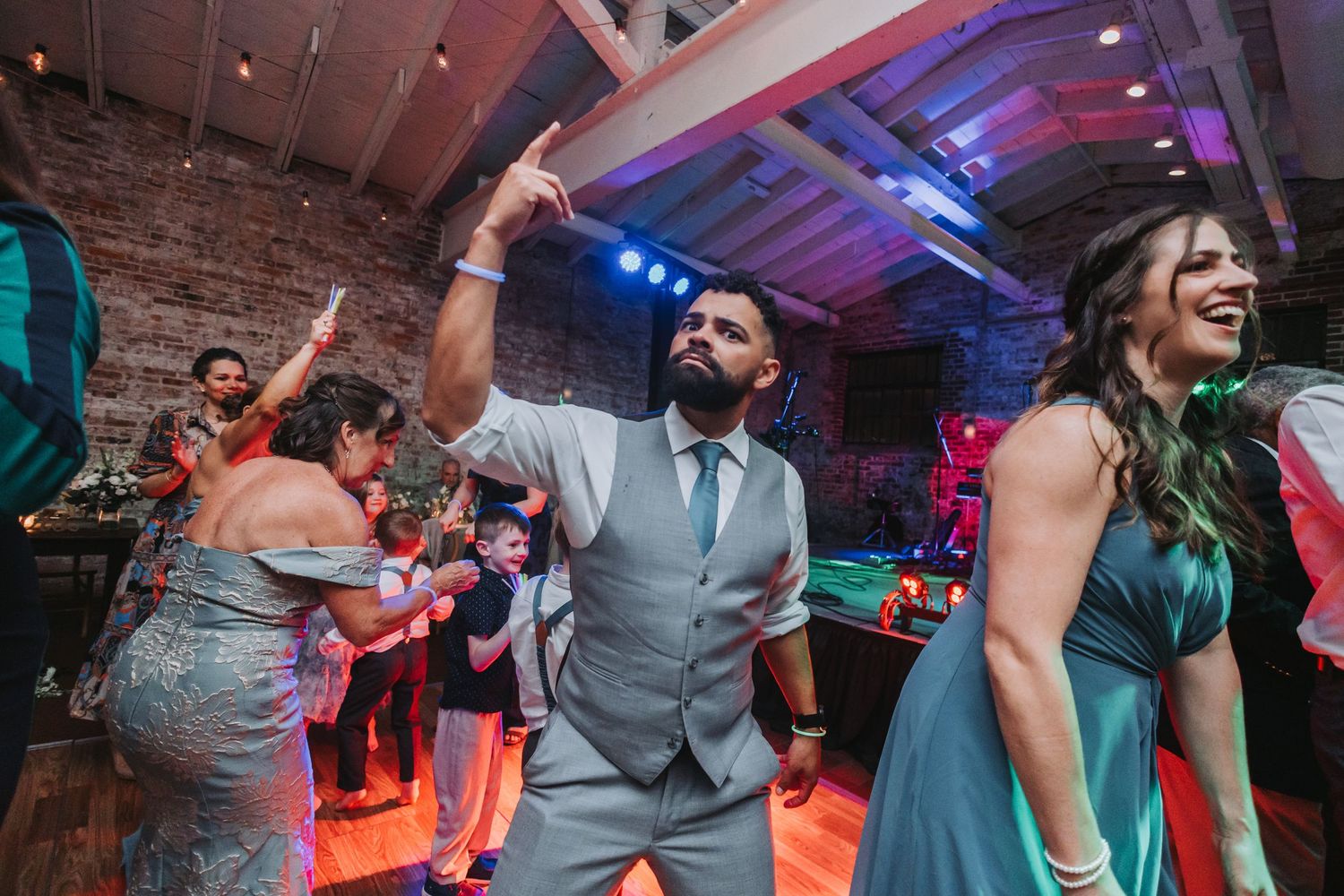 Guests dancing energetically at a wedding reception in a rustic brick venue with colorful lighting.