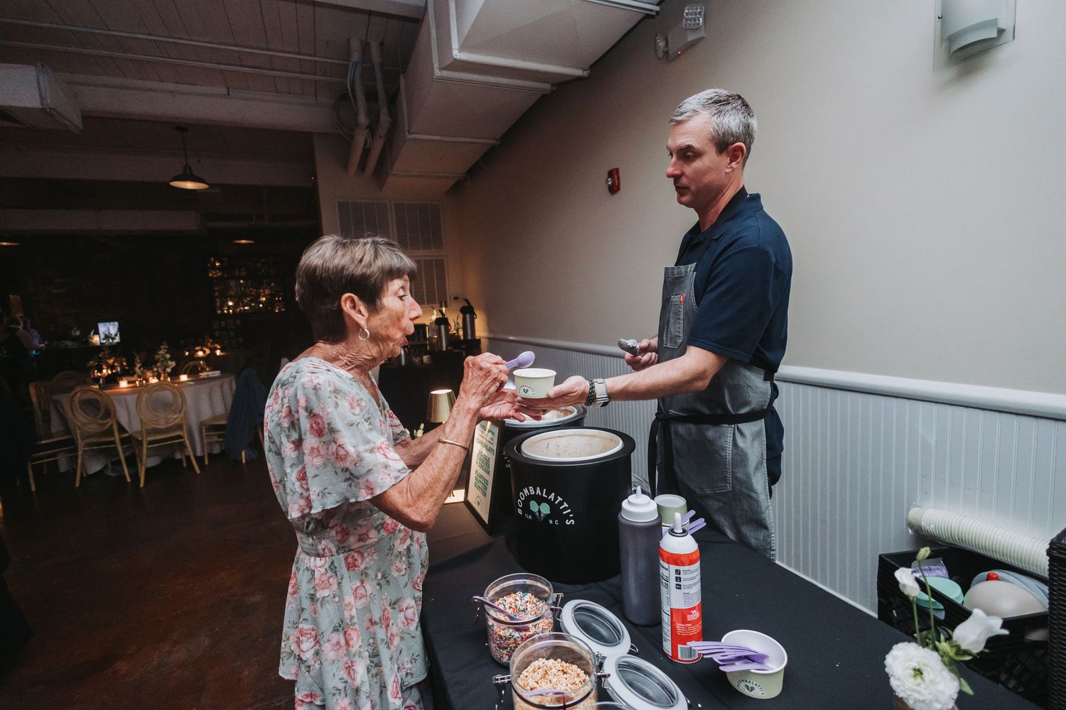 A man in an apron serves food to a woman at a catering station during an indoor event with elegant table settings.