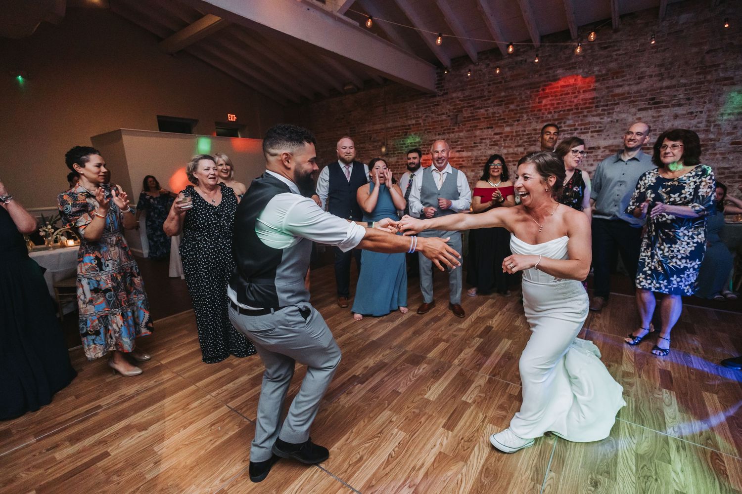 Bride and groom share a joyful first dance at wedding reception with guests clapping on wooden dance floor.