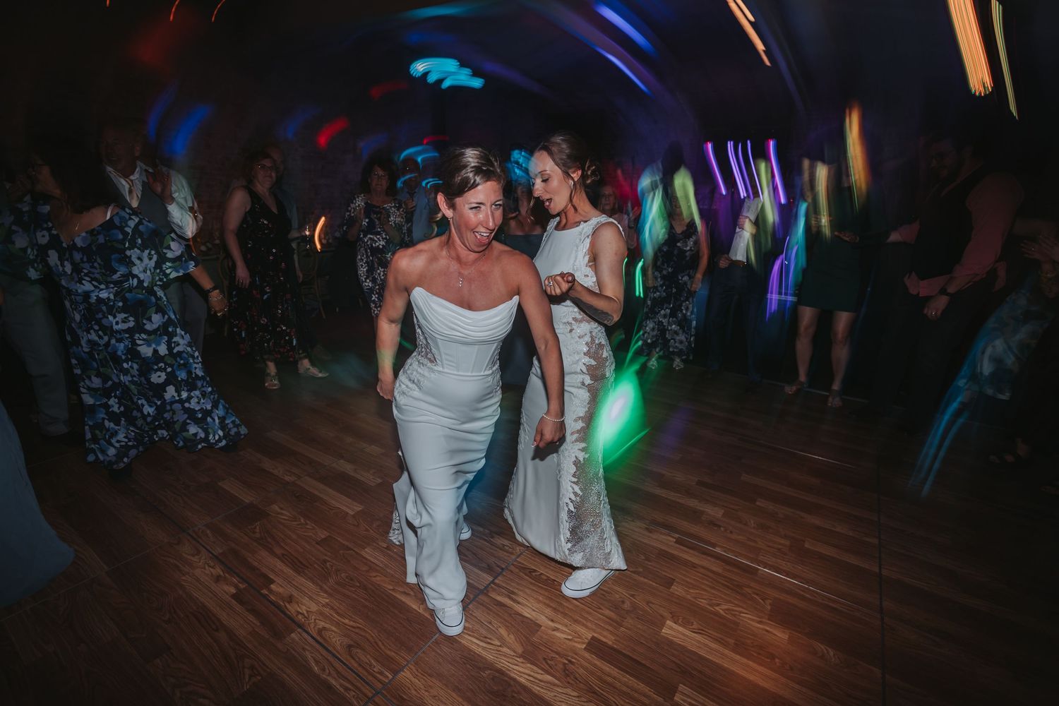 Two brides in white dresses dancing joyfully on a wooden dance floor at their wedding reception.