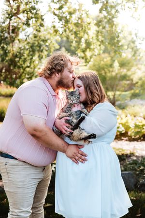 A couple kisses while holding a tabby cat outdoors in a sunny garden setting.