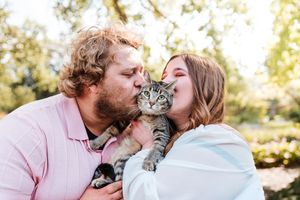 Couple kissing a tabby kitten outdoors, surrounded by green trees in a warm, sunlit park setting.
