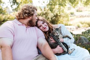 A couple cuddles outdoors with a tabby cat, the man kissing the woman's head as she holds the cat.