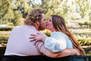Couple sharing a romantic kiss on a bench in a lush green park, woman with long hair embracing bearded man.