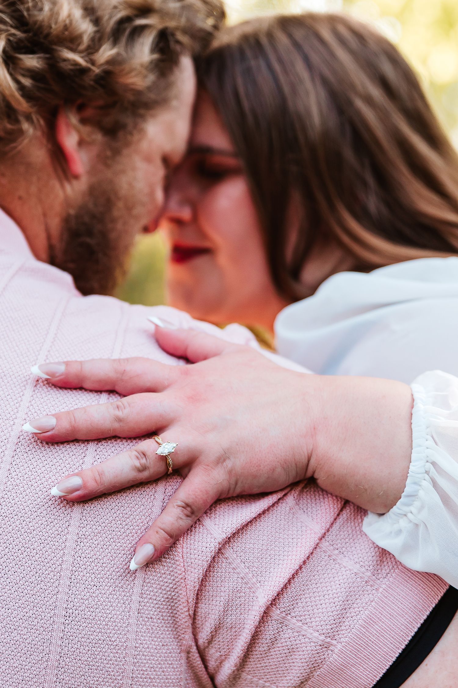 Couple embracing closely, woman's hand showing engagement ring on man's pink sweater shoulder.
