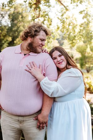 A couple embraces outdoors, man in pink polo and woman in light blue dress, smiling together in a sunlit garden setting.