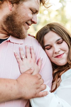 A couple shares a tender embrace outdoors, woman resting head on mans chest showing engagement ring, wearing pink and white.