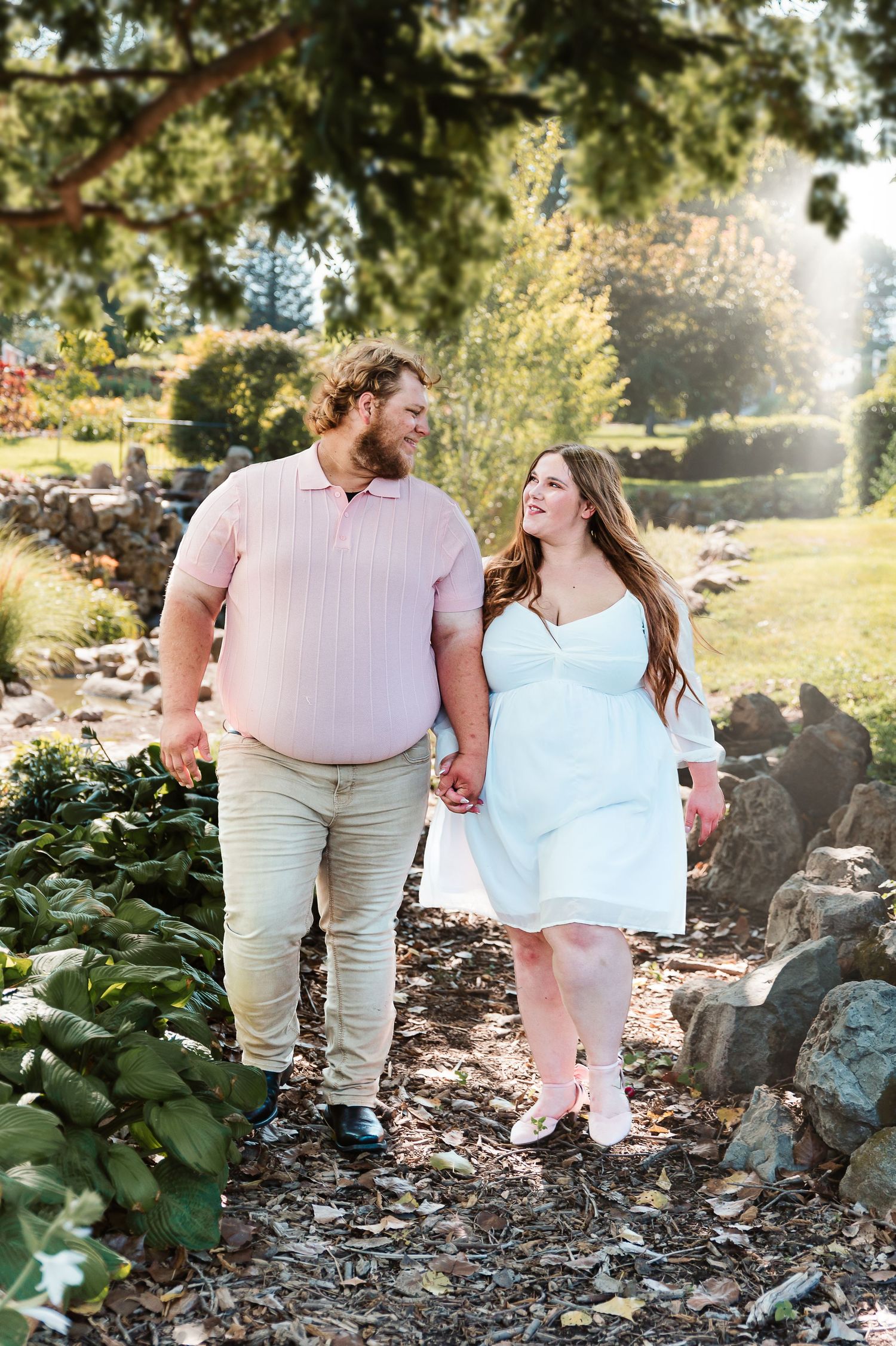 A couple holds hands and smiles at each other while walking through a sunny garden park.