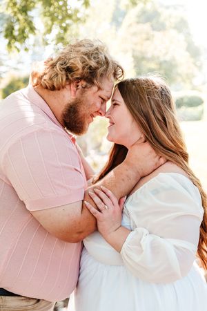 A couple in pink and white outfits share a tender forehead touch outdoors in warm sunlight.