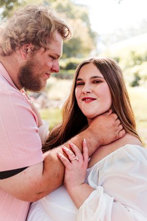 Couple in outdoor engagement photo, man in pink shirt embraces smiling woman in white dress, sunny park background.