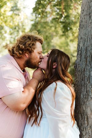 Couple sharing a romantic kiss outdoors near a tree, man in pink suit and woman in white dress.
