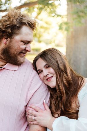 A couple shares a tender moment outdoors, woman resting head on man's chest wearing an engagement ring, surrounded by trees.