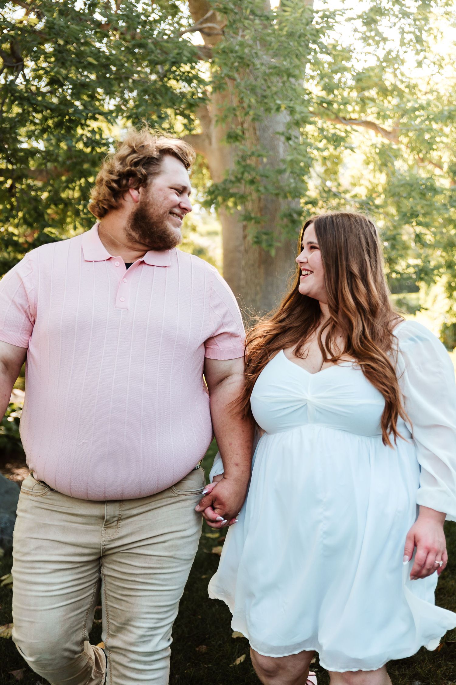 A happy plus-size couple holds hands and smiles at each other outdoors, man in pink polo, woman in white dress.