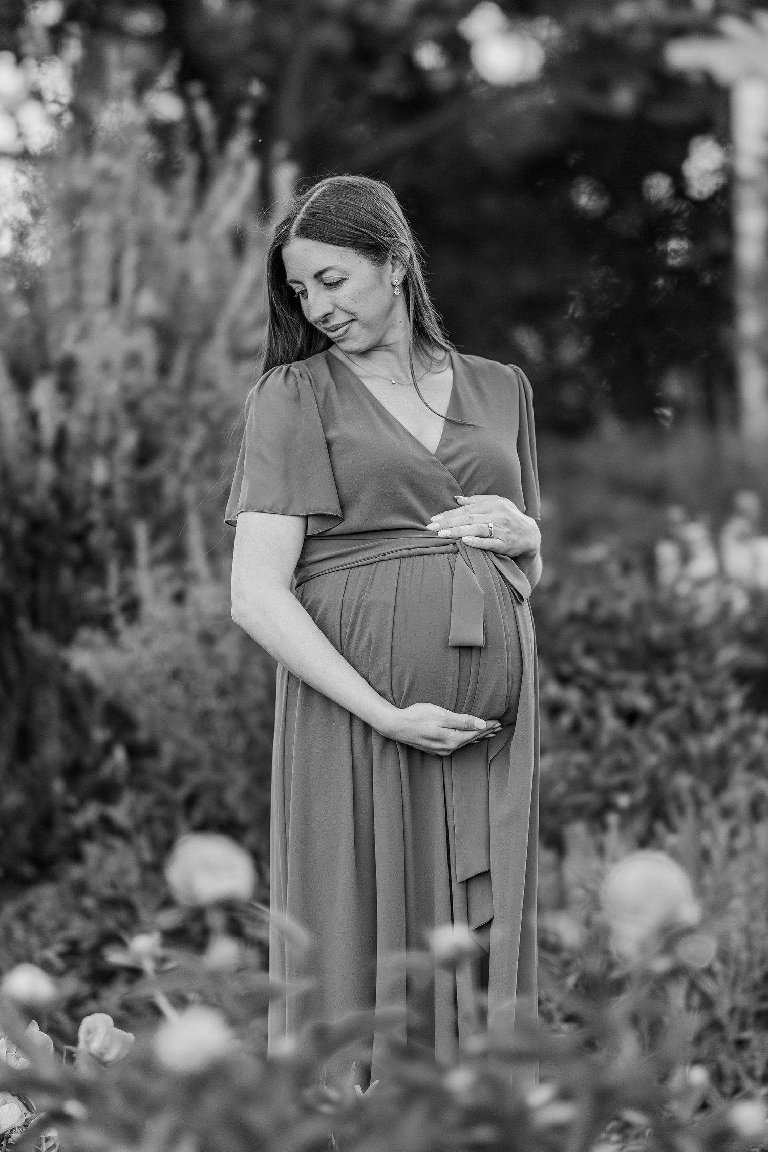 Smiling pregnant woman in a wrap dress cradling her baby bump outdoors in a garden, black and white photo.