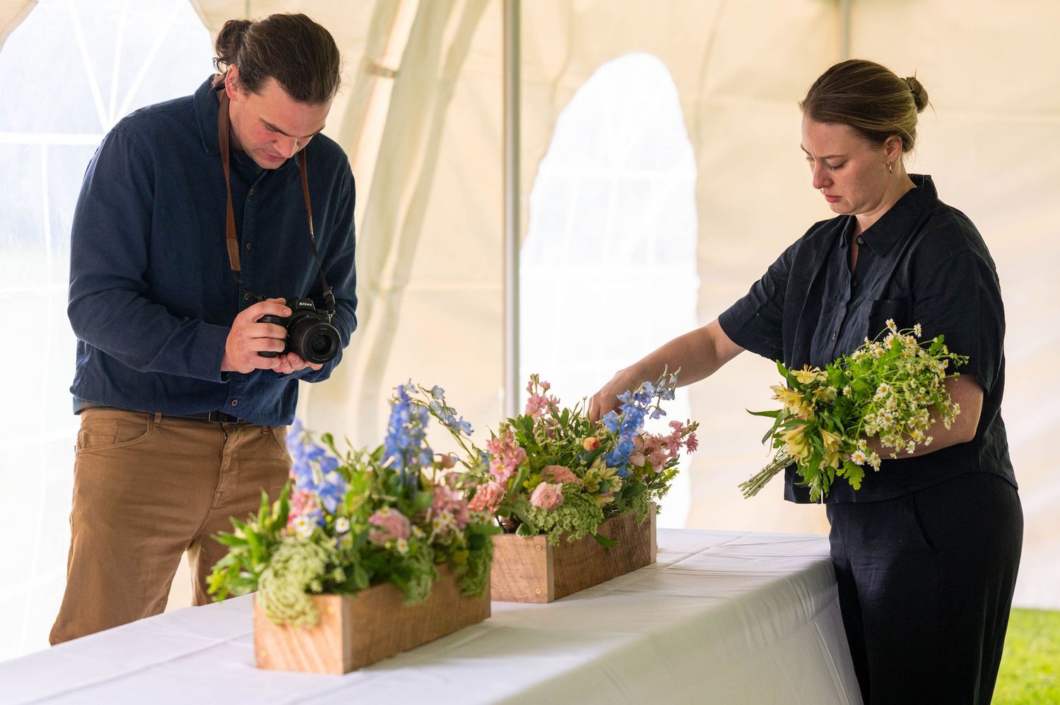 Photographer capturing floral arrangements while florist arranges colorful flowers in wooden boxes under a white tent.