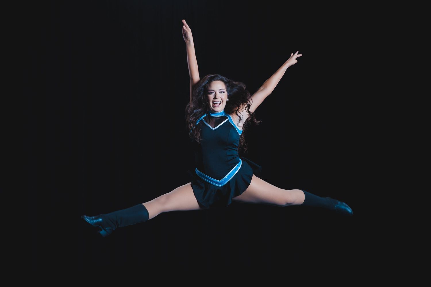 Cheerleader in black and blue uniform doing a mid-air split jump against a dark background.
