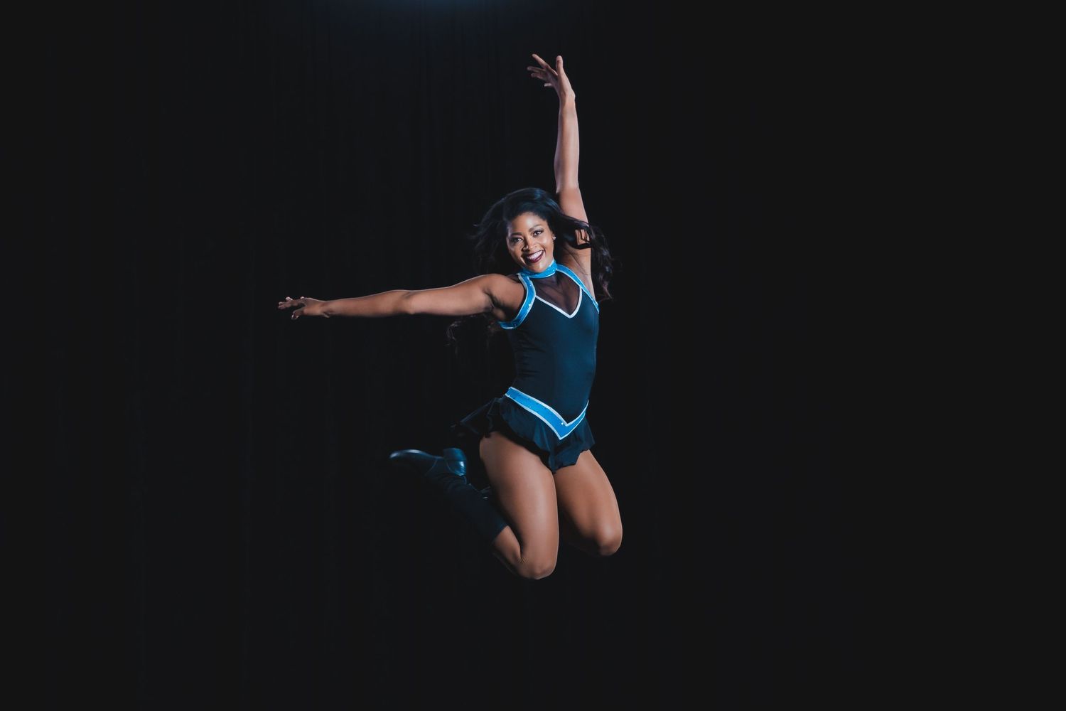 Female cheerleader in blue and black uniform jumping mid-air against dark background.