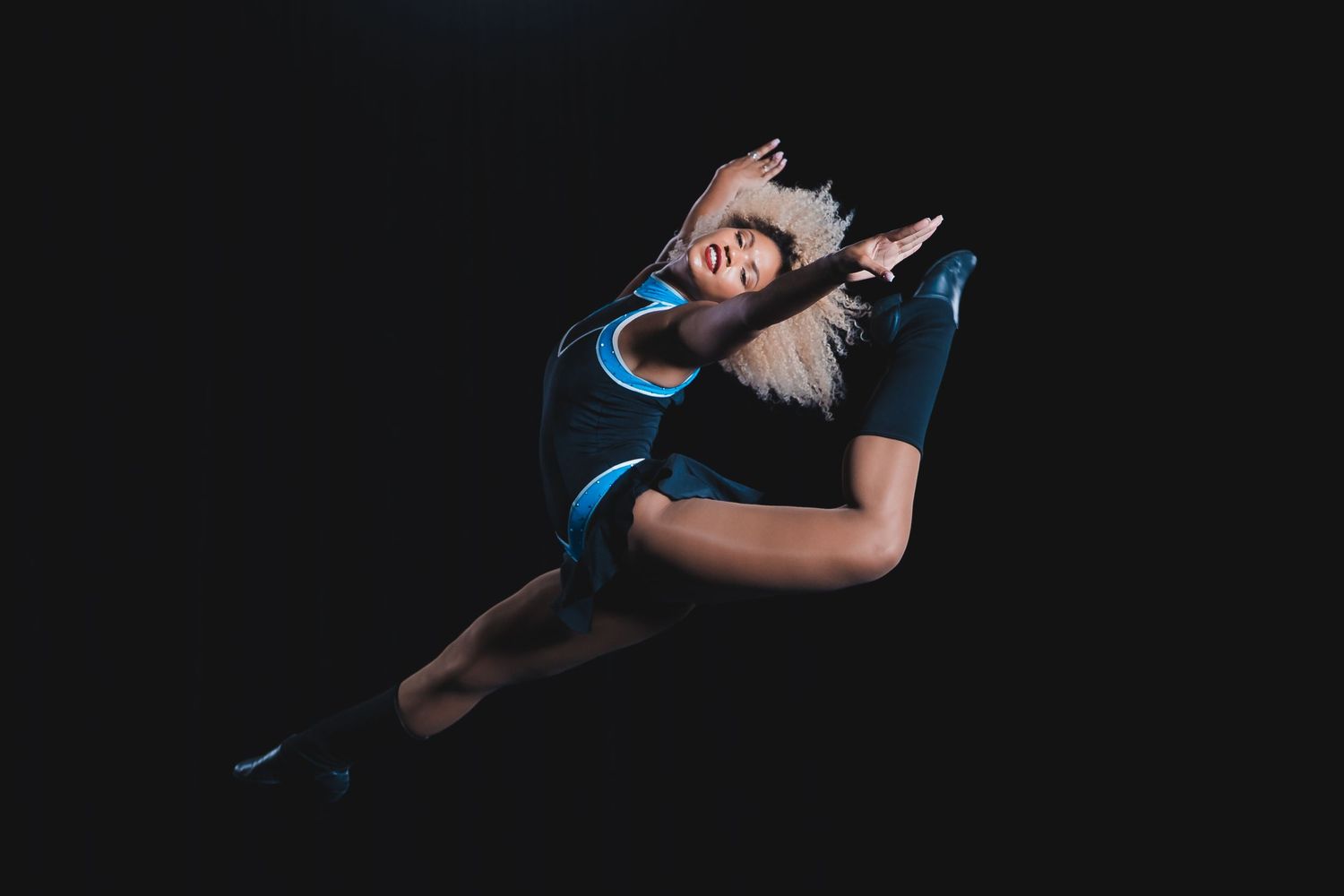 Dancer in blue costume performs a dramatic split leap against a dark background wearing a feathered headpiece.