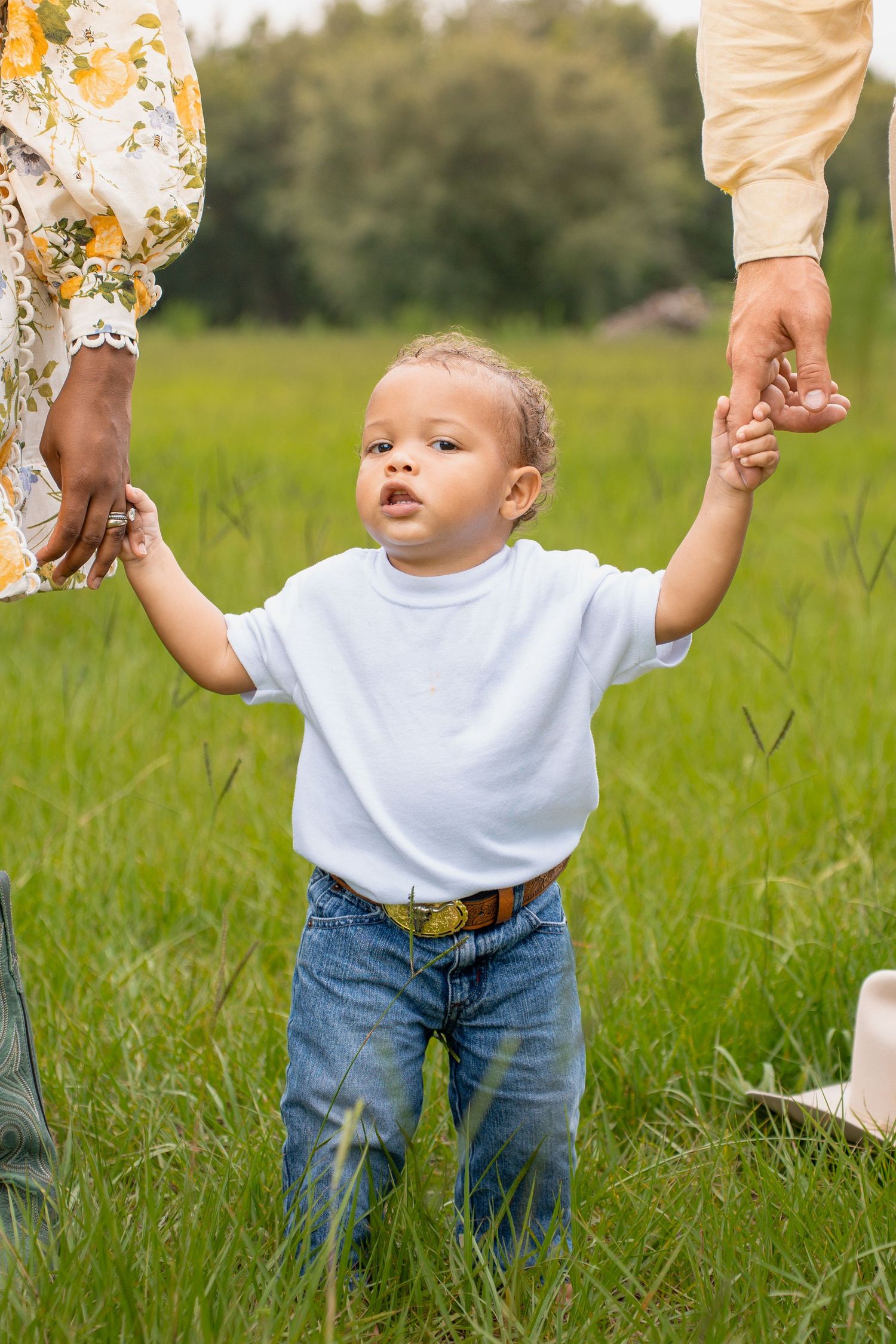Interracial couple holding hands in a green field, woman in floral dress with boots, man in cowboy hat and yellow shirt.