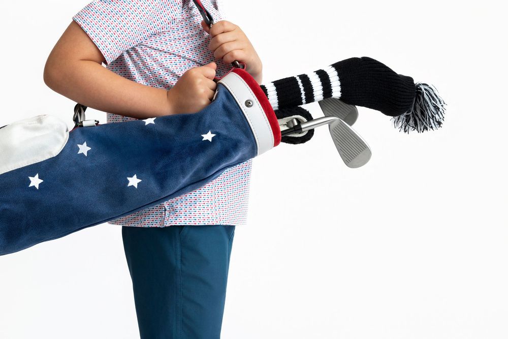 Child carrying a navy star-print golf bag with clubs and black knit headcovers on white background.