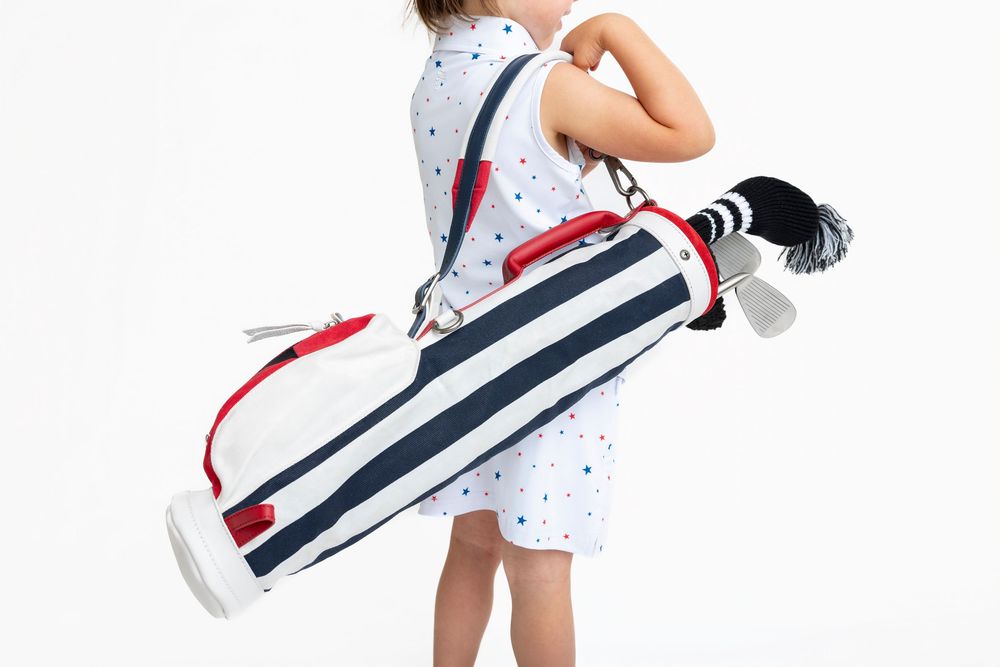 Child carrying a navy and white striped golf bag with red accents and clubs, wearing a polka dot dress.