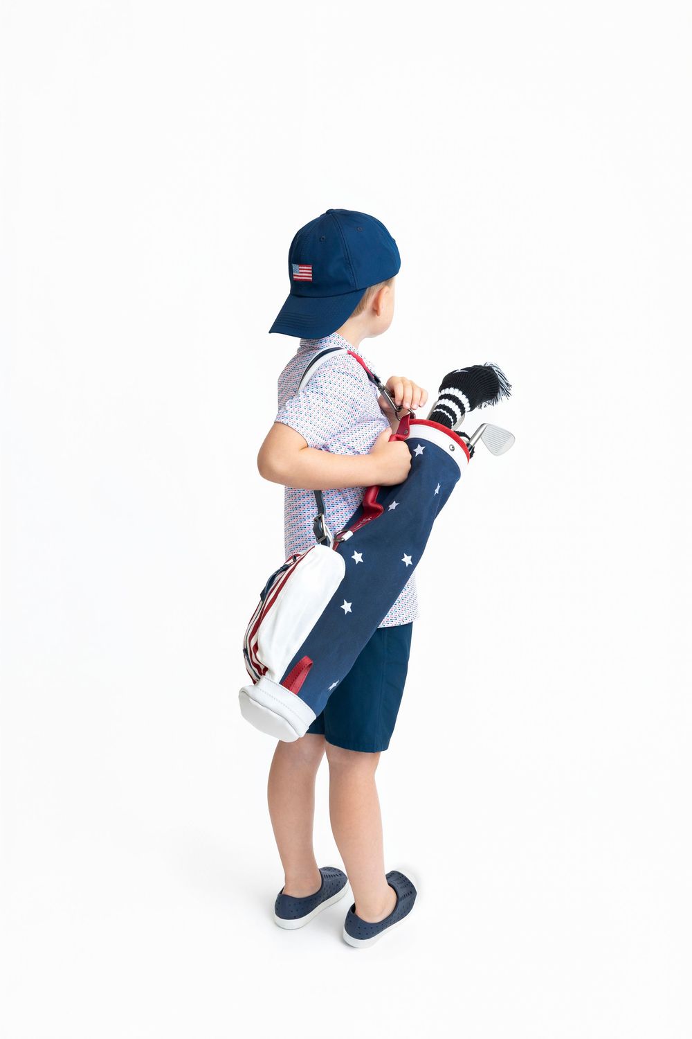 Young boy carrying a patriotic star-print golf bag with clubs, wearing navy shorts, cap, and plaid shirt.