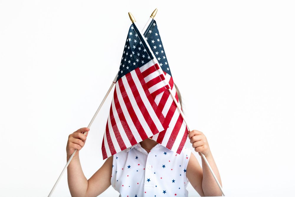 Child holding two American flags covering their face, wearing a star-patterned shirt on white background.