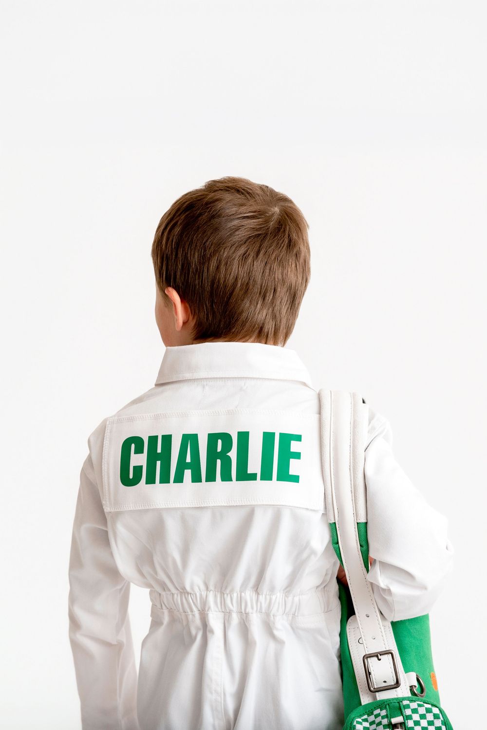 Boy wearing white jacket with CHARLIE printed on back, holding green and white checkered bag.