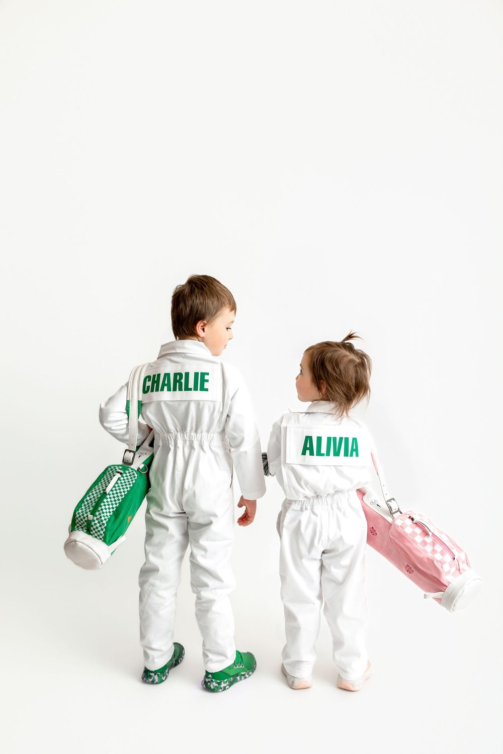 Two children in white jumpsuits named Charlie and Alivia holding colorful bags, viewed from behind.