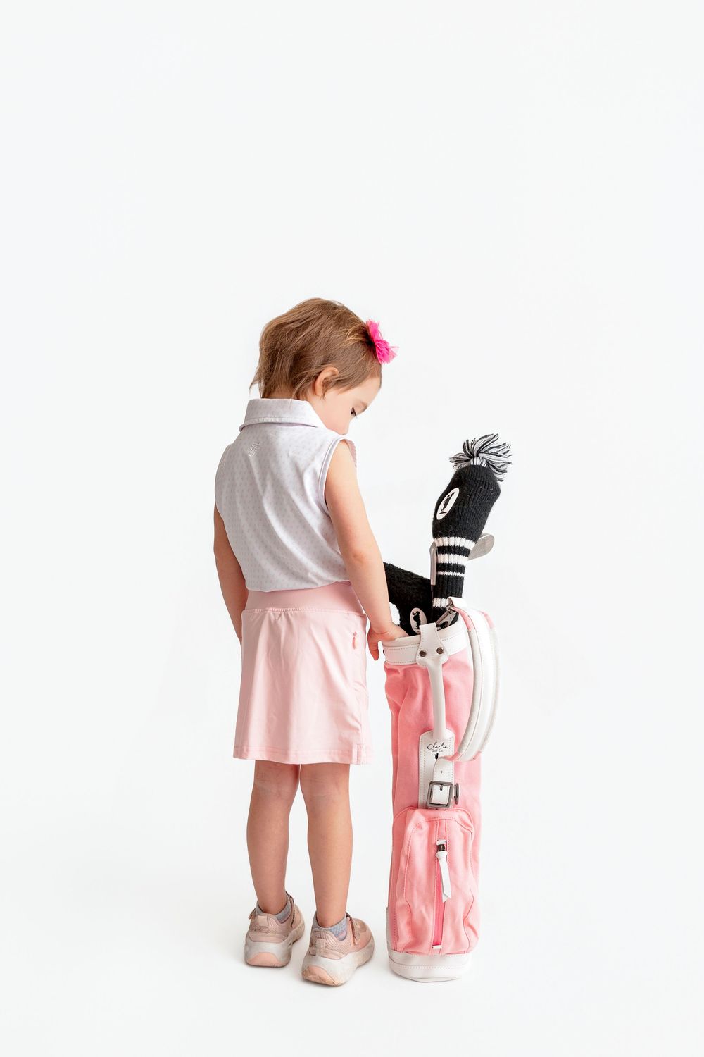 Toddler girl in white polo and pink skirt stands with a pink golf bag featuring a knitted club cover.