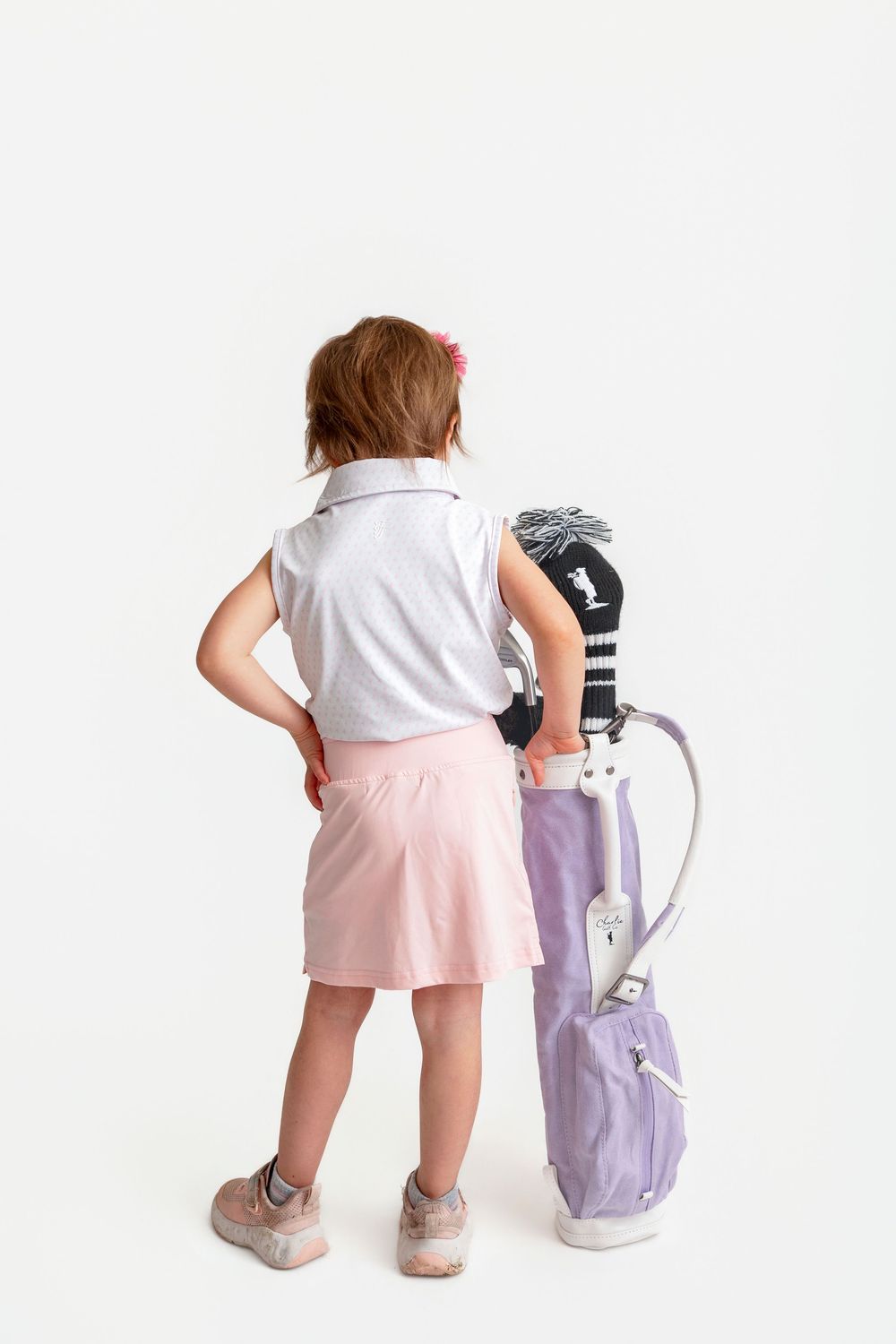 Young girl in pink skirt and white polo stands with a purple junior golf bag on a white background.