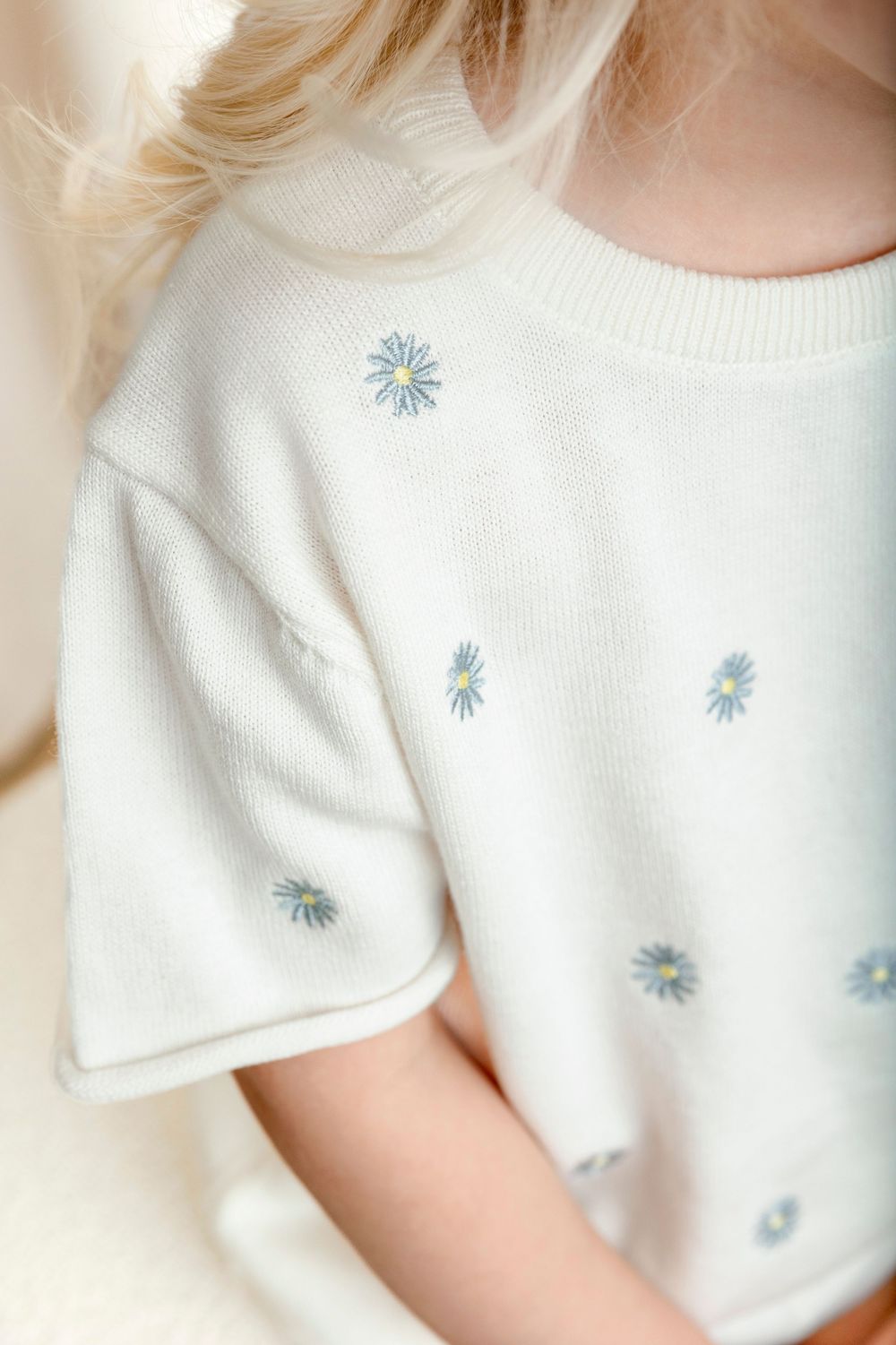 Child wearing a cream knit short-sleeve top with blue daisy embroidery pattern.