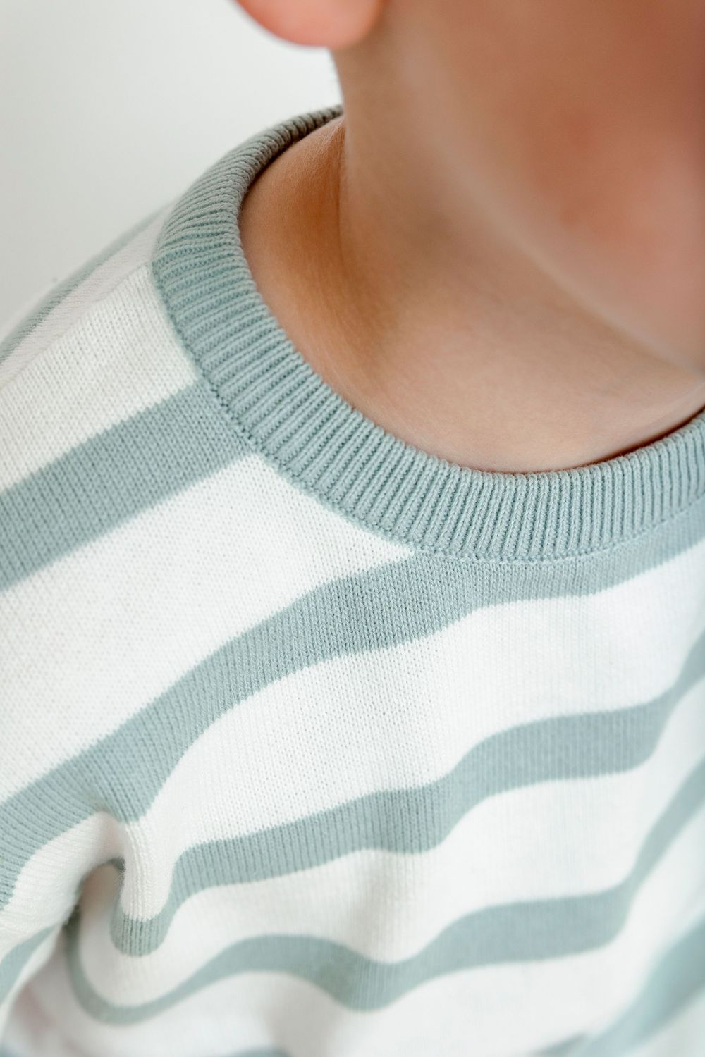 Close-up of a child wearing a sage green and white striped crew neck sweater with ribbed collar.