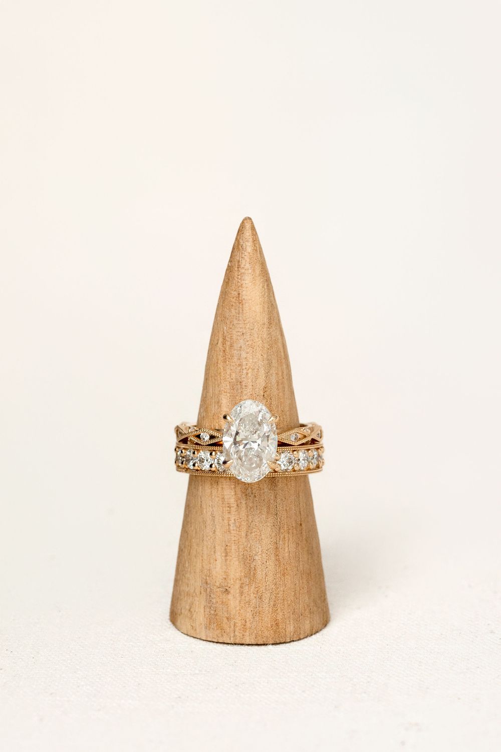 Gold engagement ring set with oval diamond solitaire and diamond band displayed on wooden cone ring holder.