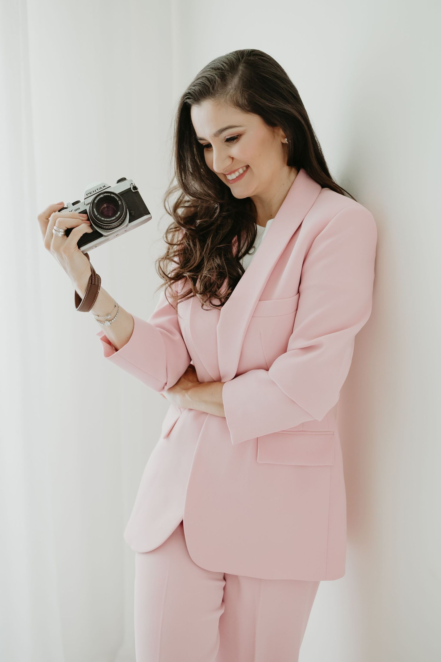 Smiling woman in pink suit holding a vintage film camera, posing against a white wall.