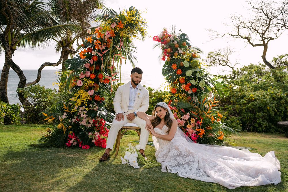 Bride and groom pose before vibrant tropical floral arch on lush green lawn at outdoor wedding ceremony.