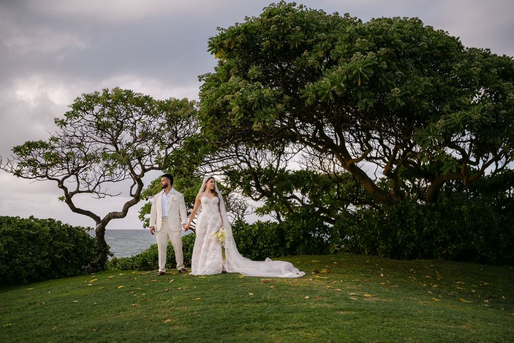 Bride and groom stand on lush green lawn surrounded by tropical trees with ocean views at outdoor wedding.