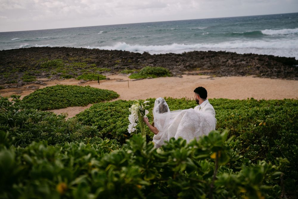 Couple in wedding attire posing on a rocky beach with lush greenery and ocean waves in the background.