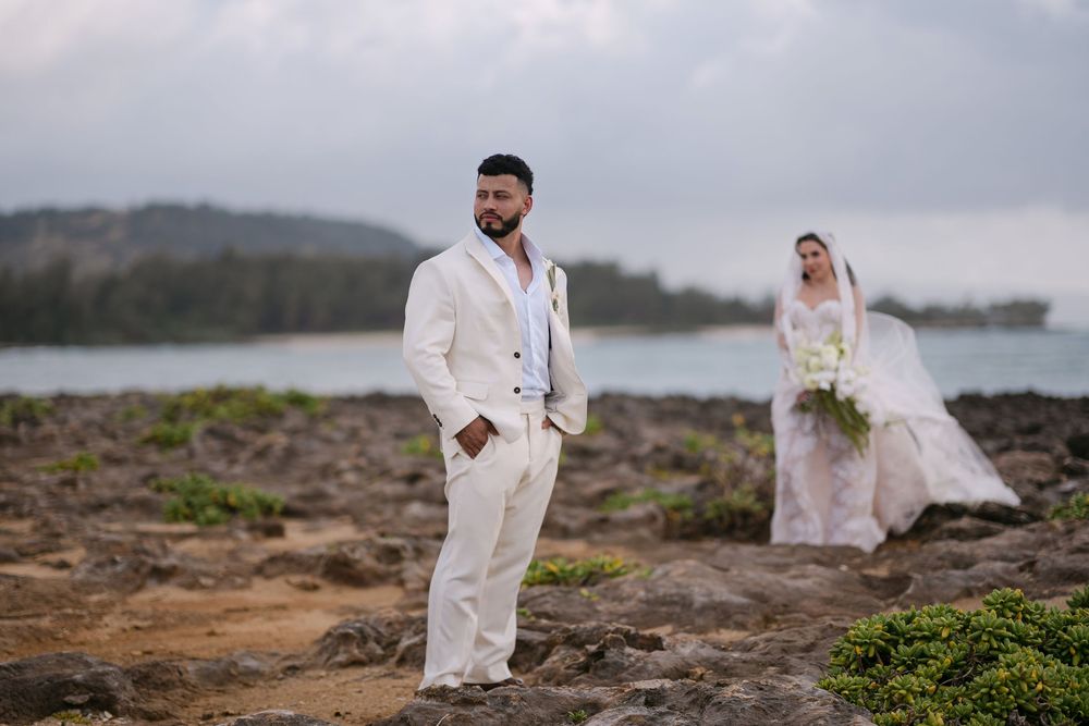 Groom in white suit stands on rocky shore with bride in flowing gown holding bouquet in scenic coastal wedding photo.