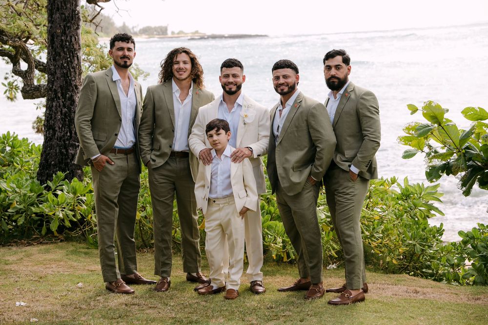Six men in olive suits pose outdoors near ocean, including a young boy in tan suit, at a tropical wedding.