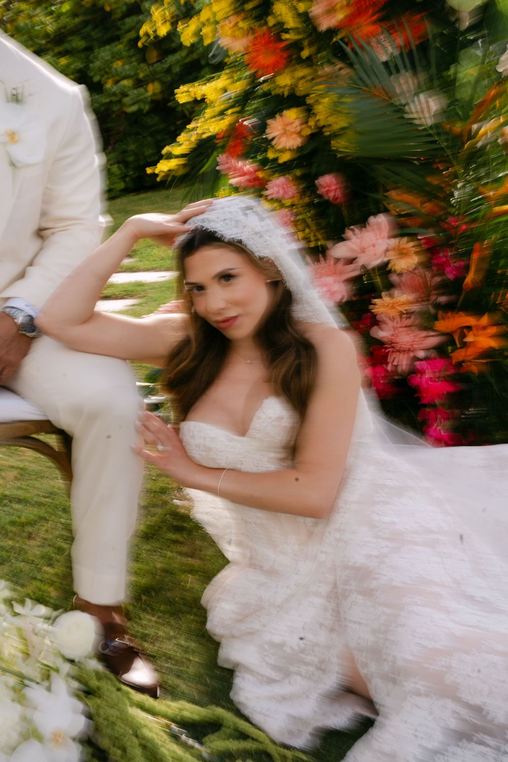 Bride in strapless white gown and veil sits gracefully outdoors beside groom, surrounded by vibrant colorful floral arrangements.