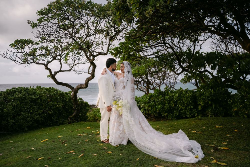 Bride and groom share a romantic kiss on lush green lawn with ocean views and tropical trees at outdoor wedding.