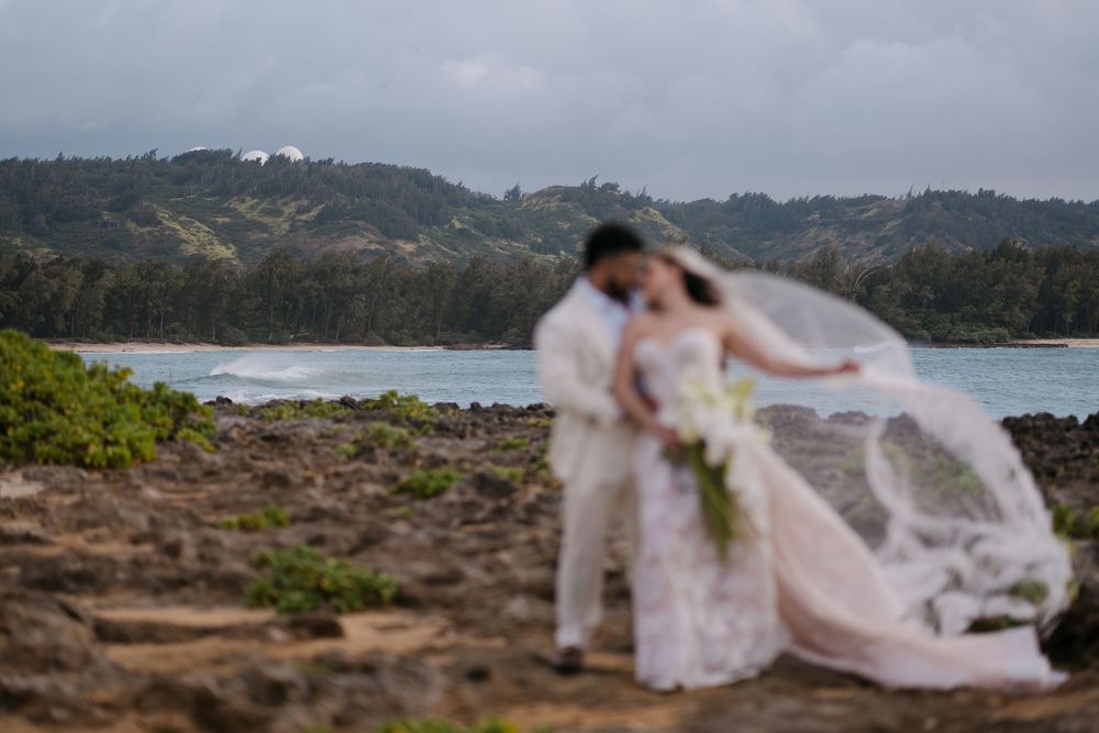 Couple shares romantic kiss in wedding attire on rocky Hawaiian coastline with lush green hills and ocean waves in background.