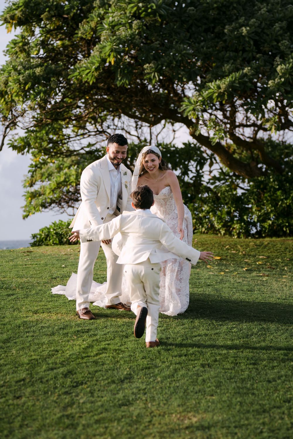 A joyful wedding couple in white attire on a lush lawn as a child runs toward them under a tropical tree.