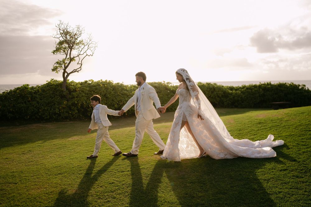 Bride and groom in white wedding attire walk hand-in-hand with a child on a lush green lawn at sunset.