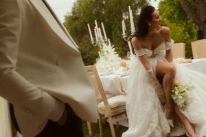 Bride in white off-shoulder gown sits at elegant outdoor wedding reception table with candles and floral arrangements.