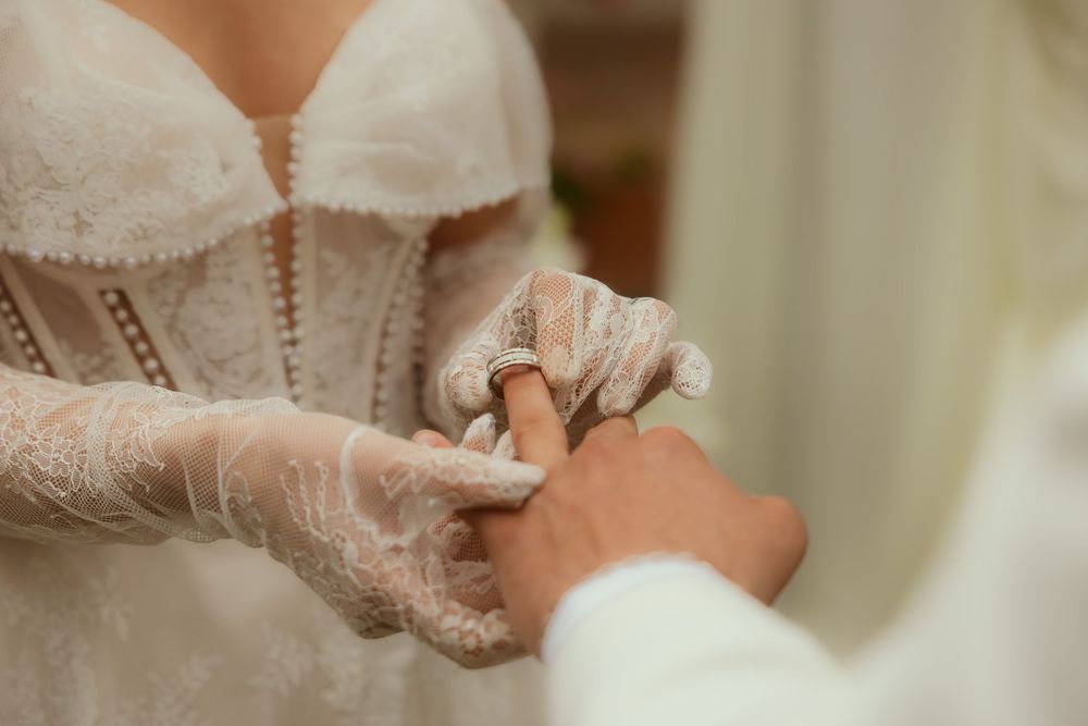Bride in lace gloves and white gown placing a wedding ring on groom's finger during ceremony.